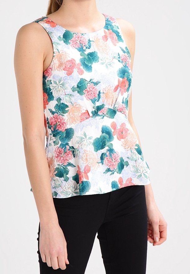 Ärmelloses Top mit floralem Muster auf weißem Hintergrund, ausgestattet mit grünen, rosa und orangefarbenen Blumen sowie einem ausgestellt geschnittenen Saum. Kombiniert mit schwarzen Hosen.