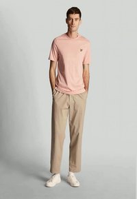 Young man standing with hands in pockets, wearing a light pink t-shirt, beige pants, and white sneakers against a plain grey background.