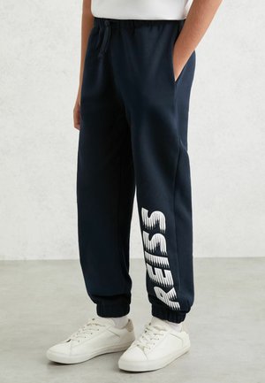 Osoba ubrana w granatowe spodnie dresowe z białymi sneakersami, ręce w kieszeniach, na lewej nodze pionowo nadrukowany biały napis "RE 313".