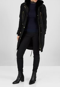 Černý dlouhý kabát se zlatými zipy a stahovacím šňůrkem, s kapucí z umělé kožešiny. Nošený přes námořnicky modrý top a černé přiléhavé kalhoty, doplněný kotníkovými botami.