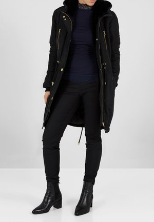 Cappotto lungo nero con cerniere dorate e coulisse, con cappuccio in pelliccia sintetica. Indossato sopra una maglia blu navy e pantaloni aderenti neri, abbinato a stivaletti alla caviglia.