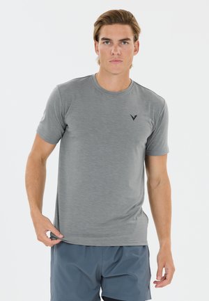 Ung mand med kort brunt hår iført en grå t-shirt med korte ærmer og blå shorts, der holder i kanten af sin skjorte.