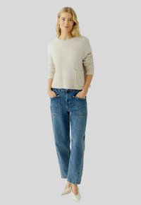Helles beiges Strickpullover mit einem gerippten Saum und einer Tasche, kombiniert mit blauen Denim-Jeans im lässigen Schnitt, die über aufgesetzte Fronttaschen und helle Nähte verfügen.