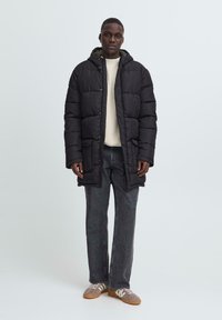 Persona con una larga chaqueta de plumas negra sobre un suéter blanco, jeans oscuros y zapatillas beige, de pie frente a un fondo claro y liso.
