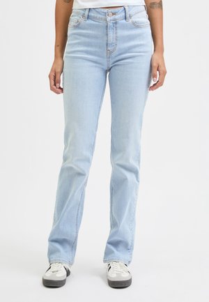 Personne portant un jean bleu clair droit et des baskets blanches, debout devant un fond blanc uni.
