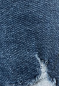 Modrá denimová tkanina s mírným umytím, obsahující poškozenou oblast s třepícími se okraji. Textura je hladká s drobnými variacemi v barvě.