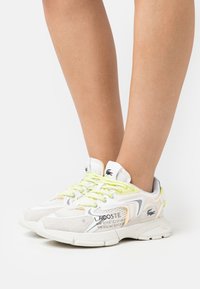 Lacoste sneakers with a mixed material design in white and light gray. Features yellow laces, textured accents, and logo detail on the side.