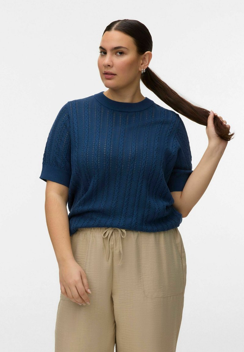 Femme aux longs cheveux foncés attachés en queue de cheval, portant un haut en maille bleu à manches courtes et un pantalon beige à cordon, debout devant un fond blanc uni.