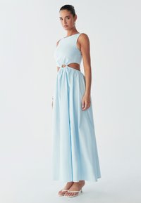 Modèle portant une robe maxi bleu clair avec un détail découpé à la taille et des sandales blanches à lanières, posée devant un fond uni.