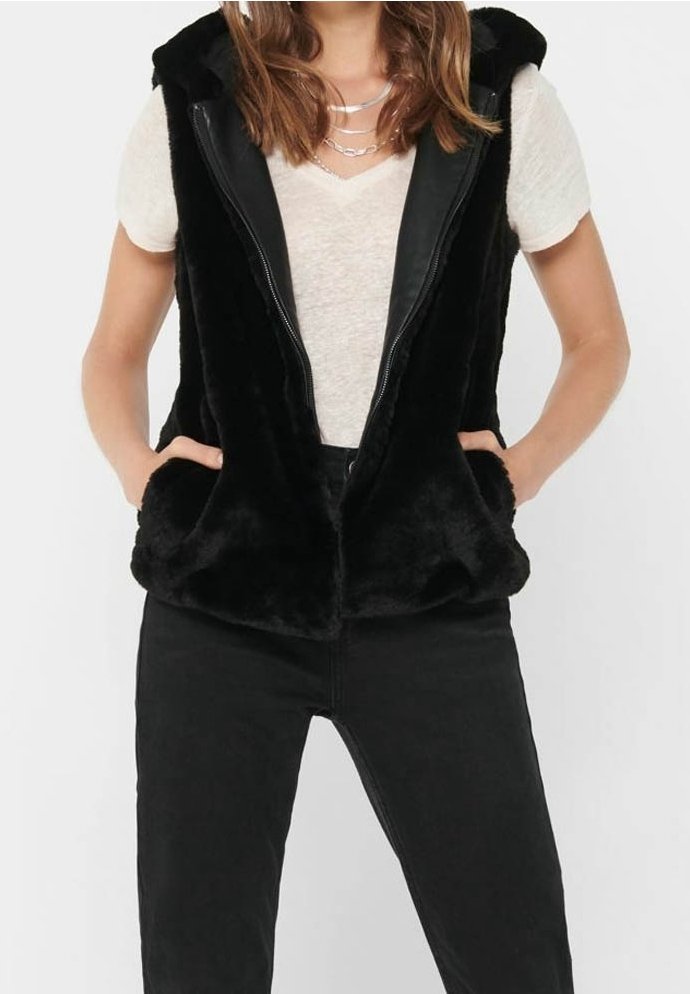 Personne portant un gilet sans manches noir en tissu duveteux avec fermeture éclair sur une chemise blanche à manches courtes, les mains dans les poches du gilet et un pantalon noir.
