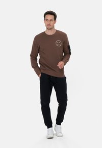 Bruine sweater met een ronde hals en witte logo-detail, gecombineerd met zwarte joggers. Model draagt witte sneakers.
