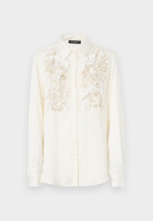 Krémová košeľa s textúrovanou kvetinovou výšivkou cez hruď, s klasickým golierom a zapínaním na gombíky. Dlhé rukávy.
