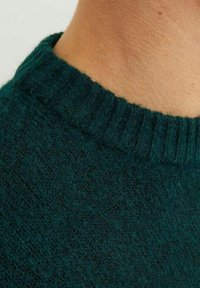 Mörkgrön stickad tröja med ribbad halsringning. Texturen ser mjuk och lurvig ut, med ett subtilt, ojämnt mönster.