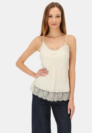 Beżowa koszulka na cienkich ramiączkach z koronkową lamówką i luźnym krojem. Posiada falistą koronkową krawędź i miękką fakturę tkaniny.