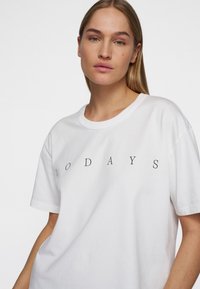 T-shirt bianco in cotone a maniche corte, con il testo "10 DAYS" in nero, centrato sul petto. Collo a giroclassico con una vestibilità rilassata.