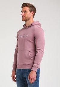 Lyserød hoodie lavet af blødt stof med en frontlomme, snøre i hætten og prægede logo på brystet. Båret med blå jeans.