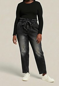 Personne portant un haut noir à manches longues, un jean noir délavé taille haute avec un cordon, et des baskets blanches, debout devant un fond uni.