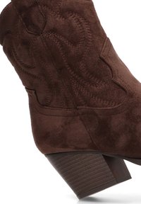 Bota de tobillo de ante marrón con un acabado texturizado, detalle de costura curvada y tacón de madera apilada. La vista lateral destaca la forma y el diseño de la bota.