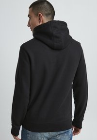 Felpa nera con un grande cappuccio a doppio strato, polsini e orlo a costine. Il materiale sembra morbido e con una consistenza liscia. Vista posteriore mostrata.