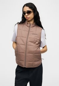 Γυναίκα με μακριά σκουρόχρωμα μαλλιά και γυαλιά ηλίου, φοράει μωβ γιλέκο με ραμμένες καπιτονέ ραφές πάνω από λευκό μπλουζάκι και μαύρο παντελόνι, μπροστά από ένα λευκό φόντο.