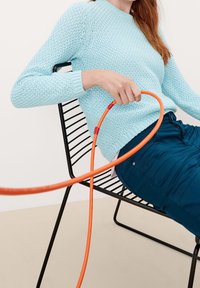Pull tricoté bleu clair avec un motif texturé, associé à un pantalon bleu foncé. Un câble en spirale orange entoure la silhouette assise.