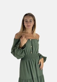Off-shoulder, olivengrøn kjole med lange, puffede ærmer. Har en smock-dekoreret talje og knapper ned foran, lavet af letvægtsstof.