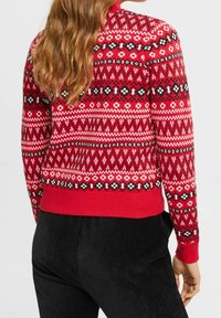 Pull tricoté rouge avec des motifs géométriques noirs et blancs, doté d'un col et d'un ourlet côtelés. Porté avec un pantalon en velours côtelé noir.