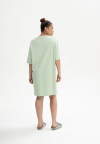 Hellgrünes, oversized Kleid aus weichem Stoff mit kurzen Ärmeln und Rundhalsausschnitt, knielanger Schnitt, getragen mit grauen Sandalen.