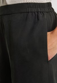 Schwarze Hose mit elastischem Bund und glatter Textur, ausgestattet mit Seitentaschen und dezenten Nähten. Nahaufnahme des Stoffs und der Tasche.