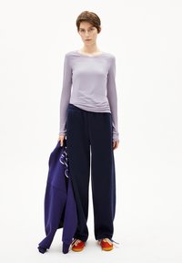 Junge Person, die in marineblauen Hosen, einem lilafarbenen Langarmoberteil und roten Turnschuhen steht und einen lila Sweatshirt mit weißem Druck auf einem weißen Hintergrund hält.