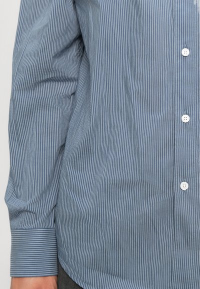Chemise bleu à manches longues avec de fines rayures verticales blanches, boutons blancs et poignets à manches retenus, portée par une personne.