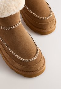 Bottes en suédine marron avec un manchon en fausse fourrure blanche, ornées de coutures décoratives et d'une semelle en caoutchouc. Bout arrondi avec une texture douce.