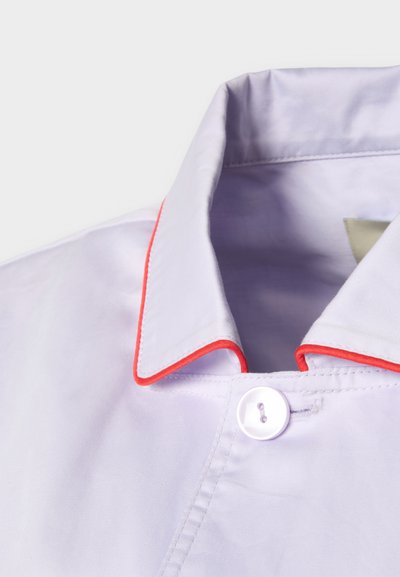 Κοντινό πλάνο για το γιακά ενός ανοιχτό μωβ πουκαμίσου με κόκκινη διακόσμηση και λευκό κουμπί σε απλό φόντο.