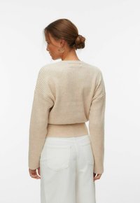 Femme aux cheveux bruns attachés en chignon, portant des boucles d'oreilles créoles argentées, un pull côtelé beige et un pantalon blanc, vue de dos sur un fond blanc.