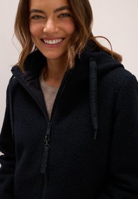 Zwarte fleece jas met een ritssluiting en capuchon. Beschikt over een textuurstof, verstelbare trekkoorden en een logo op de ritssluiting en capuchon.