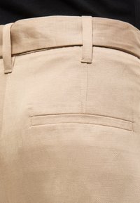 Close-up af beige bukser med en baglomme med klap og bæltestropper, båret med en sort top.