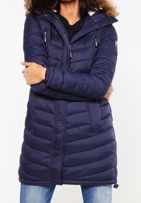 Marinblå vadderad kappa med luva fodrad med päls, utrustad med fickor med dragkedja, insvängd midja och ett strukturerat quiltmönster.