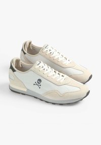 Baskets avec une tige en toile crème et en daim, accents gris, icône de squelette noire, lacets blancs et semelle en caoutchouc texturée.