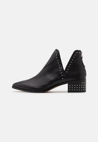 Botte cheville en cuir noir avec un bout pointu, des côtés ouverts et une fermeture éclair. Talon épais orné de clous en argent. Texture lisse.