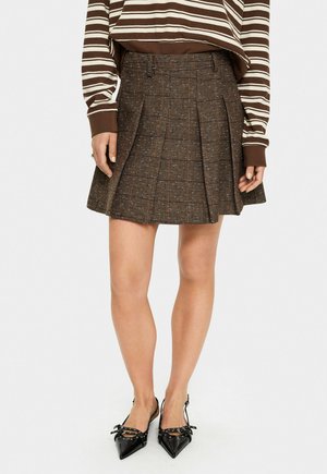 Brauner Tweed-Faltenrock mit Karomuster. Texturiert mit dezenten blauen Punkten. Kombiniert mit schwarzen spitzen Schuhen.