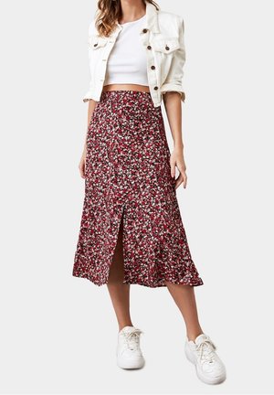Woman wearing white cropped jacket, white crop top, red floral midi skirt with front slit, and white sneakers standing against light background.