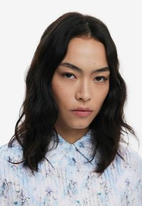 Light blue, pleated blouse with a floral pattern. Features a pointed collar and three blue buttons along the front. Smooth fabric texture.