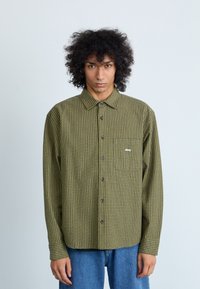 Groene geruite button-up shirt met een borstzak, bruine knopen en een klein "obey" logo. Lange mouwen met knoopmanchetten. Spijkerbroek.