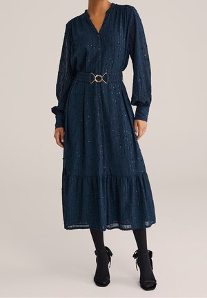 Granatowa sukienka midi z długim rękawem wykonana z fakturowanej tkaniny, z przodu zapięta na guziki, z paskiem ze złotą klamrą podkreślającym talię, noszona z czarnymi rajstopami i butami na obcasie.