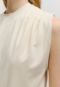 Beige mouwloos topje met een ronde halslijn, vervaardigd van zachte, gladde stof en met een fraaie draping bij de schouders.