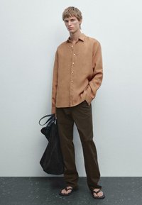 Chemise en lin marron à manches longues, coupe décontractée, avec des boutons. Pantalons en coton marron foncé, portant un sac en tissu noir, et portant des tongs noires.