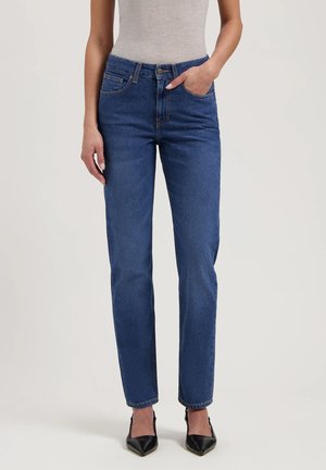 Woman wearing blue straight-leg jeans with five pockets and black pointed-toe heels, standing against a plain background.