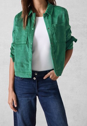 Kobieta w jasnym zielonym zapinanym na guziki płaszczu na białej bluzce, ubrana w ciemnoniebieskie dżinsy, stojąca z jedną ręką w kieszeni.