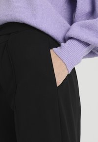 Světle fialový pletený svetr s žebrovanými manžetami, v kombinaci s černými elegantními kalhotami s bočními kapsami. Důraz na texturu a barevný kontrast.