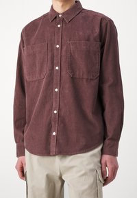 Camisa de pana en burdeos, con cuello abotonado, dos bolsillos en el pecho y un corte relajado, con tejido texturizado visible y botones blancos.